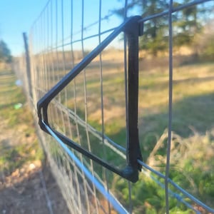 Op de afbeelding: Een zwarte plastic hek-poortvergrendeling bevestigd aan een draadhek. De vergrendeling is driehoekig en heeft een klein gat bovenaan.