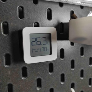 Peut inclure: Un thermomètre numérique carré blanc affichant 26,3 degrés Celsius et 70% d'humidité. L'appareil est monté sur un panneau perforé gris foncé. L'écran affiche également un petit visage souriant.