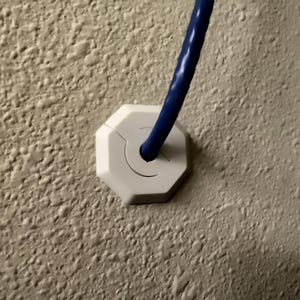Op de afbeelding: Witte plastic kabelmanagementkap met een blauwe kabel die er doorheen loopt, gemonteerd op een getextureerde muur.