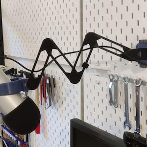 May include: A silver microphone with a black foam cover is mounted on a black, wave-shaped arm. The arm is attached to a white pegboard wall. Tools hang below the pegboard.