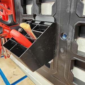 May include: A black plastic tool organiser with several compartments is mounted on a wall. A red tool and a paintbrush are visible inside the organiser. The organiser is part of a larger system with additional storage slots.