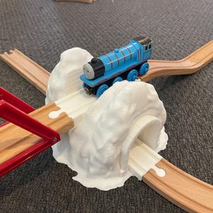 May include: A blue toy train engine with a white face and black wheels is going through a white plastic tunnel. The tunnel is attached to a wooden train track.