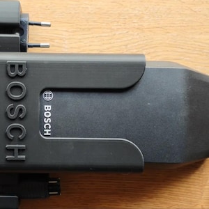 Puede incluir: Un cargador de batería Bosch negro, con el logotipo de Bosch en un lugar destacado. El cargador tiene forma rectangular con bordes redondeados y múltiples puertos de carga. Incluye un enchufe y un cable de carga.