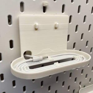 Puede incluir: Un organizador de panel perforado blanco impreso en 3D con un estante y un soporte para cables integrados. El estante contiene un cable blanco con un conector USB-C. El organizador está montado en un panel perforado blanco.