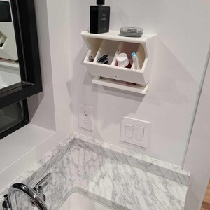 May include: A white, wall-mounted bathroom organiser with several compartments. It holds a black cologne bottle, a small container, razors, a tube of toothpaste, and other toiletries. The organiser is mounted above a marble countertop.