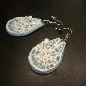 May include: A pair of silver earrings with white Millennium Falcon charms. The charms are detailed with the iconic ship's design.
