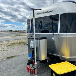 May include: A silver Airstream trailer with a satellite dish mounted on the back. The trailer is parked on a paved lot with a red and black wheel chock and a yellow plastic storage bin.