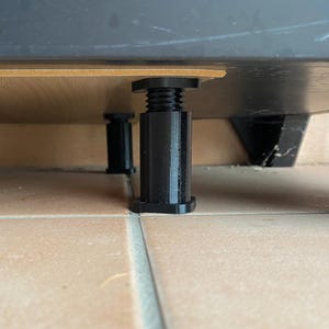 May include: Black adjustable furniture legs supporting a wooden surface. The legs have a threaded design for height adjustment and are positioned on a tiled floor. The image shows the underside of a piece of furniture.