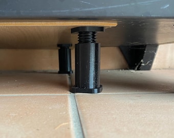 May include: Black adjustable furniture legs supporting a wooden surface. The legs have a threaded design for height adjustment and are positioned on a tiled floor. The image shows the underside of a piece of furniture.