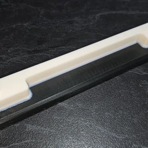May include: A white plastic handle with two raised knobs on a black base. The handle is designed to be used as a grip for a tool or object.