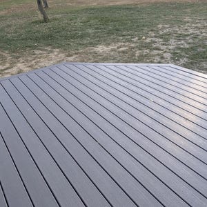 Puede incluir: Una cubierta compuesta gris con un diseño multiángulo. La cubierta está hecha de tablas largas y estrechas con una sutil textura de vetas de madera. El fondo presenta una zona de césped y algunos árboles.