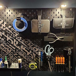 Op de afbeelding: Zwart pegboard met diverse gereedschappen en accessoires, waaronder tape, tangen, scharen en schroevendraaiers. Het bord heeft een zilver en zwart geometrisch ontwerp aan de linkerkant. Twee lampen zijn bovenop gemonteerd.