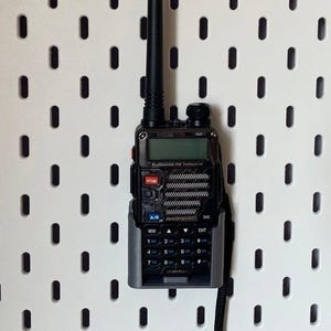 Puede incluir: Un transceptor FM profesional portátil negro y gris con pantalla y botones. La radio tiene una antena negra y está montada en una placa perforada blanca.