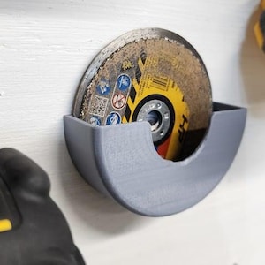 May include: A grey 3D-printed holder for a circular saw blade, mounted on a white surface. The blade is yellow and brown with safety symbols. Other tools are visible in the background.