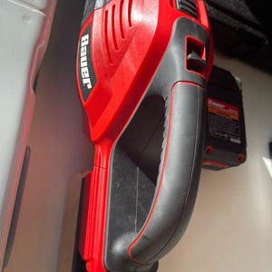 May include: A red and black Bauer impact wrench mounted on a wooden holder. The tool has a black handle and a red body with the Bauer logo. A black battery pack is visible nearby.