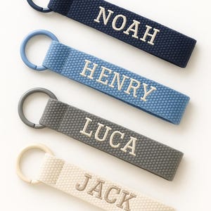 May include: Four personalized keychains in navy, light blue, gray, and off-white. Each keychain features a name embroidered in white: Noah, Henry, Luca, and Jack. Each has a matching ring.