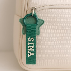 Può includere: Un portachiavi verde a forma di stella con il nome "SINA" ricamato in bianco su una cinghia verde. Il portachiavi è attaccato a uno zaino color crema con una cerniera argentata.