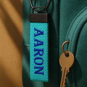 May include: A teal keychain with the name "AARON" embroidered in blue. The keychain is attached to a black metal ring and is hanging on a teal backpack. A brass key hangs next to the keychain.