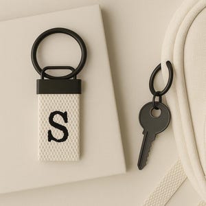 May include: A black key and a keychain with the letter "S" embroidered in black on a white woven background. The keychain has a black metal ring and a black rectangular top. The key and keychain are on a light beige surface.