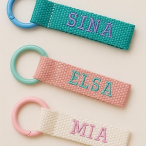 May include: Three personalised keyrings with embroidered names. The top keyring is teal with the name "SINA" in purple, the middle is pink with "ELSA" in teal, and the bottom is white with "MIA" in pink. Each has a matching coloured ring.