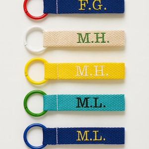 May include: Five personalised keyrings in navy blue, beige, yellow, teal, and navy blue. Each keyring has a different initial combination embroidered in a contrasting colour, with a matching ring.