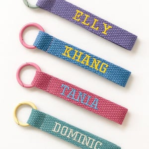 May include: Four personalized keychains in purple, blue, pink, and teal. Each keychain has a different name embroidered in a contrasting color: Elly, Khang, Tania, and Dominic. The keychains have matching colored metal rings.