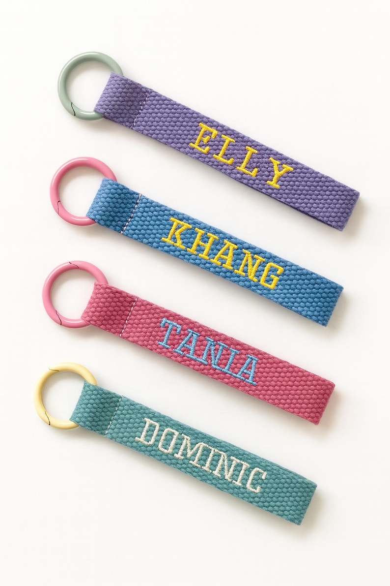May include: Four personalized keychains with embroidered names: Elly, Khang, Tania, and Dominic. Each keychain features a different color strap: purple, blue, pink, and teal. The names are embroidered in contrasting colors, with a metal ring.