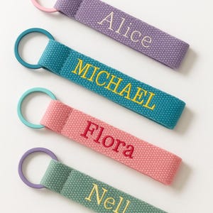 May include: Four personalized keychains in various colors: purple, teal, pink, and green. Each keychain features a different name embroidered in a contrasting color: Alice, Michael, Flora, and Nell. The keychains have matching colored rings.