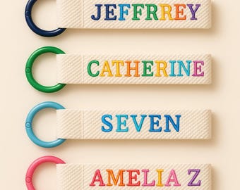 May include: Four personalized keychains with names embroidered in rainbow colors. Each keychain has a different colored ring: navy, green, light blue, and pink. The names are Jeffrey, Catherine, Seven, and Amelia Z, all on a cream-colored background.