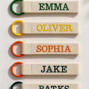May include: Five personalised keyrings with embroidered names: EMMA, OLIVER, SOPHIA, JAKE, and BATKS. Each keyring features a cream-coloured fabric strap with a coloured metal ring. The names are embroidered in green, yellow, and orange.
