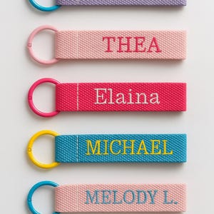 Puede incluir: Una foto vertical de cinco llaveros personalizados. Cada llavero tiene una correa de tela con un nombre bordado en un color contrastante. Los llaveros son lavanda, rosa, rojo, azul y rosa claro, con los nombres "Aloe", "THEA", "Elaina", "MICHAEL" y "MELODY L."