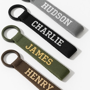 May include: Four personalized keychains in gray, black, olive green, and brown. Each keychain features a name embroidered in a contrasting color: Hudson, Charlie, James, and Henry. The keychains have a textured fabric and a metal ring.