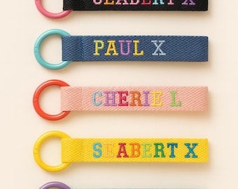 May include: Five colorful keychains with embroidered names. The keychains have a loop and a rectangular strap. The straps are black, blue, pink, yellow, and teal. The names are in various colors. The loops are pink, teal, red, yellow, and purple.