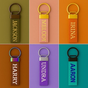Könnte beinhalten: Eine Sammlung personalisierter Schlüsselanhänger in verschiedenen Farben, jeweils mit einem anderen Namen. Die Schlüsselanhänger haben ein Stoffband mit einem Metallring und einem Metallakzent oben. Namen sind Jackson, Melody, Irina, Harry, Undra und Aaron.