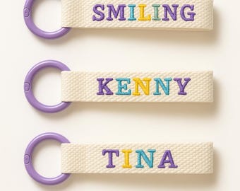 May include: Three cream-colored keychains with purple hardware. Each keychain has embroidered text in colorful letters: "SMILING", "KENNY", and "TINA". The keychains are likely for personal use or as gifts.