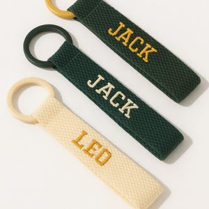 May include: Three personalized keychains. Two are dark green with "JACK" embroidered in gold, and one is cream-colored with "LEO" embroidered in gold. Each keychain has a matching ring in gold, black, and cream.
