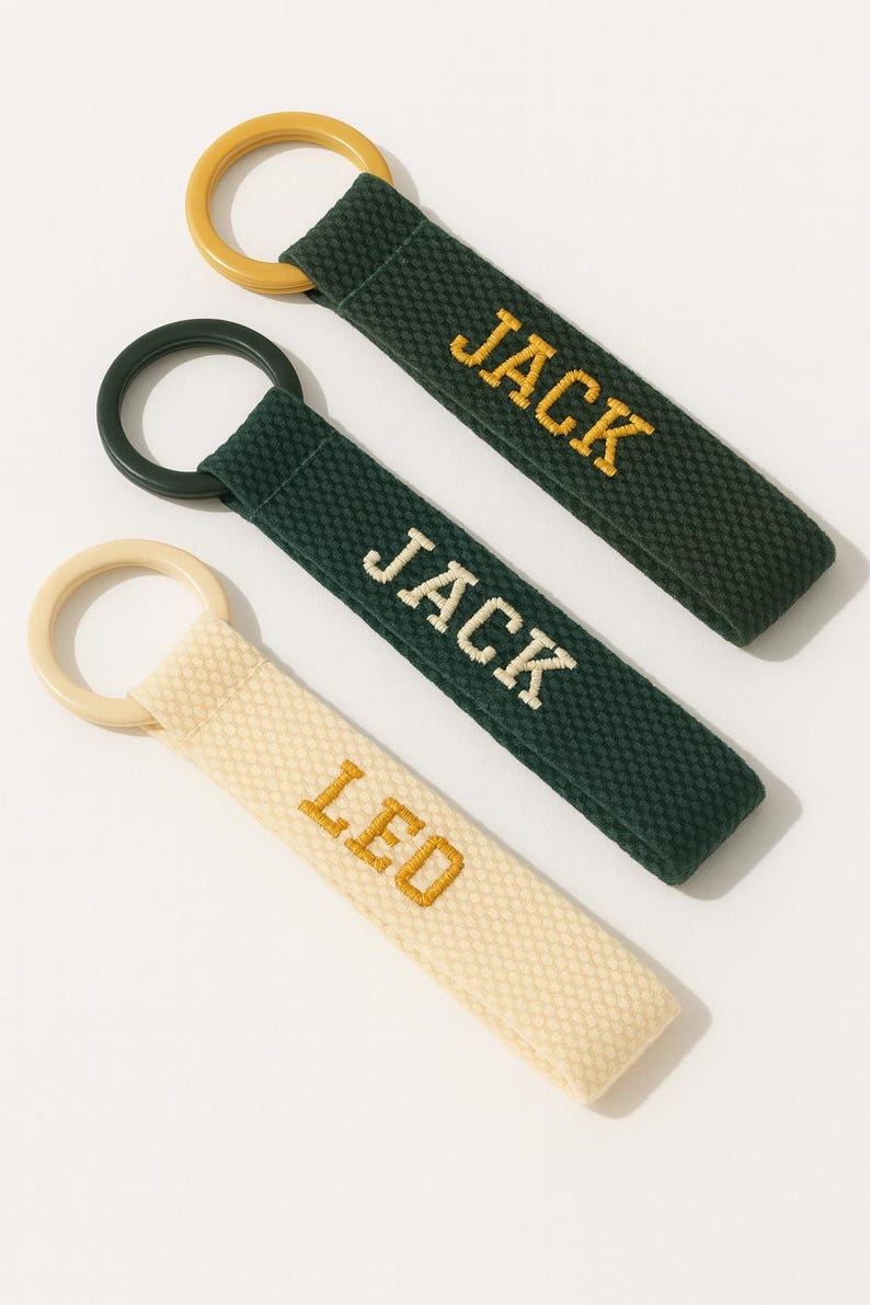 May include: Three personalized keychains with embroidered names. Two are dark green with "JACK" in gold, and one is cream-colored with "LED" in gold. Each keychain has a circular ring in a coordinating color.