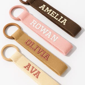 May include: Four personalized keychains with names embroidered in various colors. The keychains are in shades of brown, pink, tan, and cream, each with a matching ring. The names Amelia, Rowan, Olivia, and Ava are displayed.