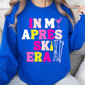 Könnte beinhalten: Ein königsblaues Sweatshirt mit den Worten "IN MY APRES SKI ERA" in rosa, weißen und gelben Blockbuchstaben. Ein rosa Cocktailglas und weiße Skistöcke sind ebenfalls auf der Vorderseite des Sweatshirts abgebildet.
