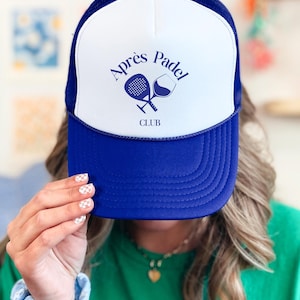 Op de afbeelding: Blauwe en witte truckerpet met de tekst "Après Padel Club" in marineblauw. Het ontwerp bevat een padelracket en een wijnglas. De pet heeft een mesh achterkant en een gebogen klep.
