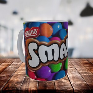 Op de afbeelding: Witte keramische mok met een kleurrijk ontwerp van Nestle Smarties snoepjes. De mok heeft het Nestle logo en het woord "Smarties" in een bruin en wit lettertype.