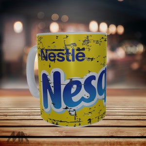 Puede incluir: Una taza de cerámica blanca con fondo amarillo y un logotipo de Nestle Nesquik azul y blanco. El logotipo tiene un aspecto desgastado y vintage.