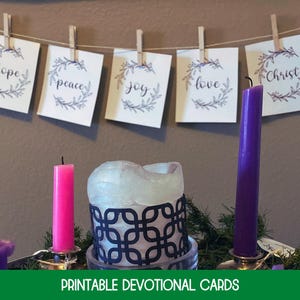 May include: Five white square cards hanging on a string with wooden clips, each featuring a botanical wreath and the words hope, peace, joy, love, and Christ. Below are three candles, one pink and two purple, with a central white candle in a dark holder.