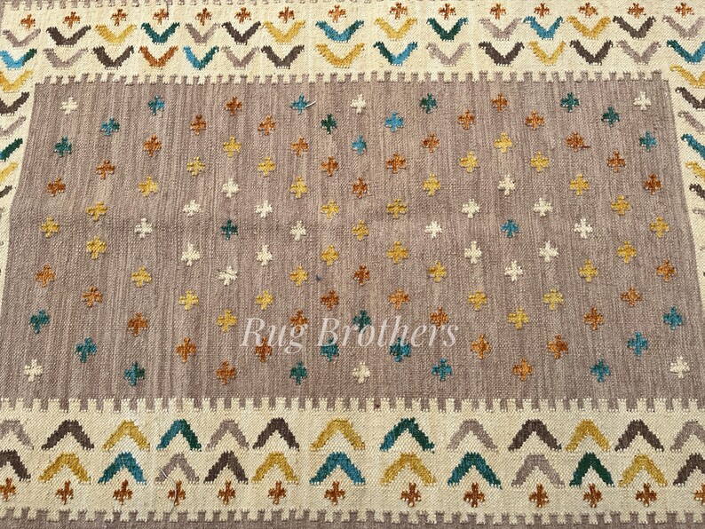 May include: A rectangular rug with a central field of muted gray, adorned with scattered cross-shaped motifs in teal, gold, and white. The border features a repeating geometric pattern in complementary colors. The text "Rug Brothers" is visible.