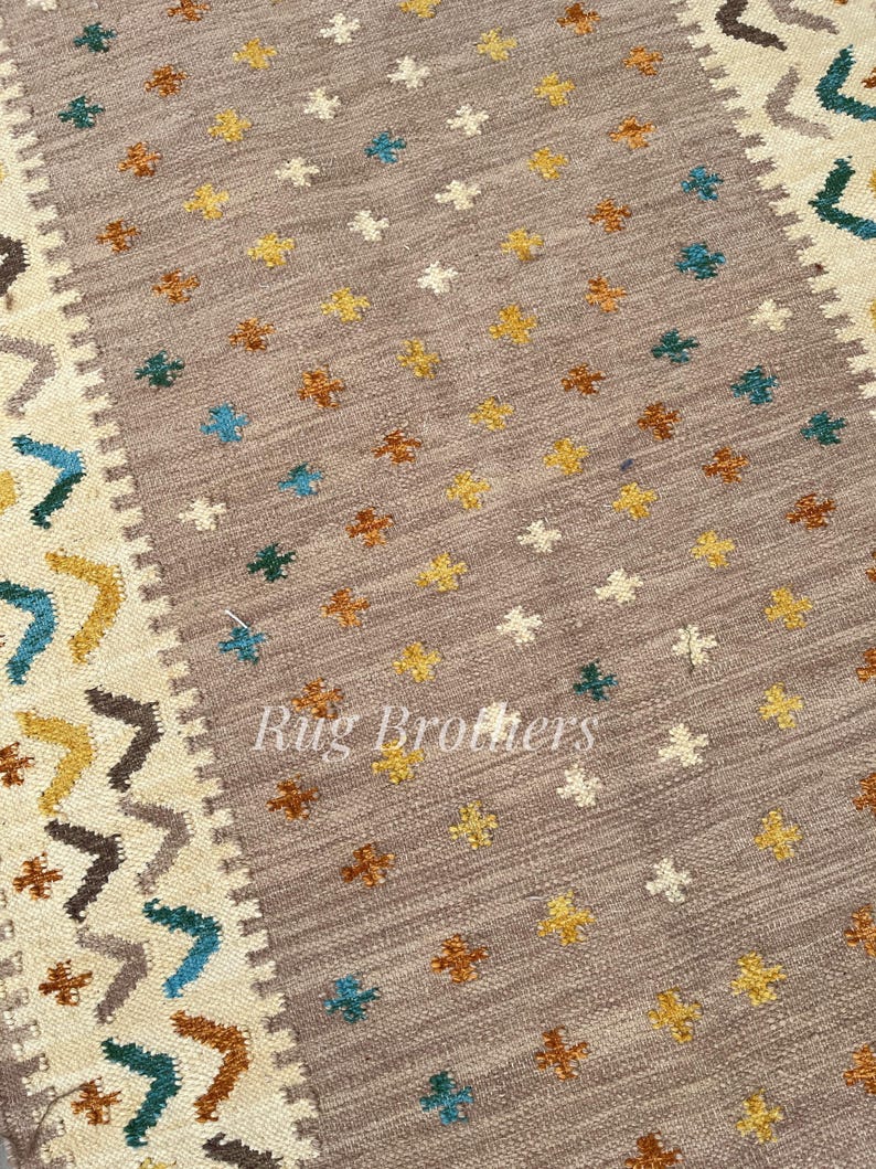 May include: A rectangular area rug with a central field of taupe, adorned with small, repeating floral patterns in yellow, brown, white, and teal. The border features geometric designs in similar colors. The text "Rug Brothers" is visible.
