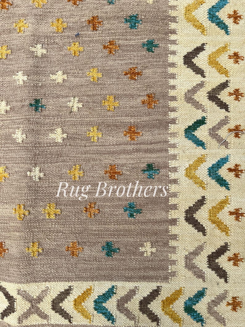 May include: Close-up of a woven rug with a neutral gray background. The rug features a pattern of small, colorful floral motifs in yellow, teal, and brown. The border has a geometric design with chevron and cross patterns. The text "Rug Brothers" is visible.