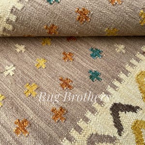 May include: Close-up of a rolled-up, textured rug with a muted mauve background. The rug features small, colorful floral patterns in yellow, orange, and teal. The text "Rug Brothers" is visible.