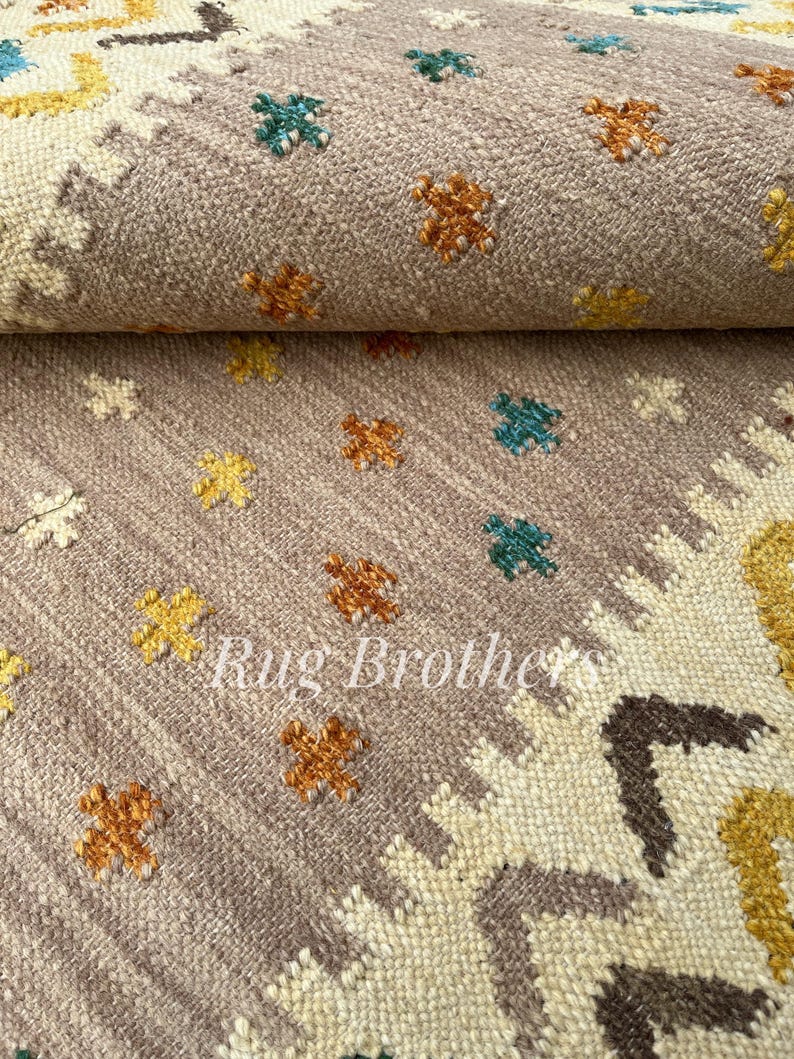 May include: Close-up of a rolled-up, textured rug with a muted mauve background. The rug features small, colorful floral patterns in yellow, orange, and teal. The text "Rug Brothers" is visible.