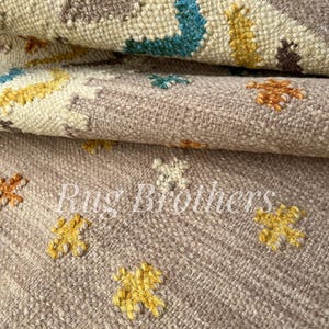 May include: Close-up of a beige and brown woven rug with a repeating floral pattern in yellow, blue, and orange. The rug has a textured surface and the text "Rug Brothers" is visible.
