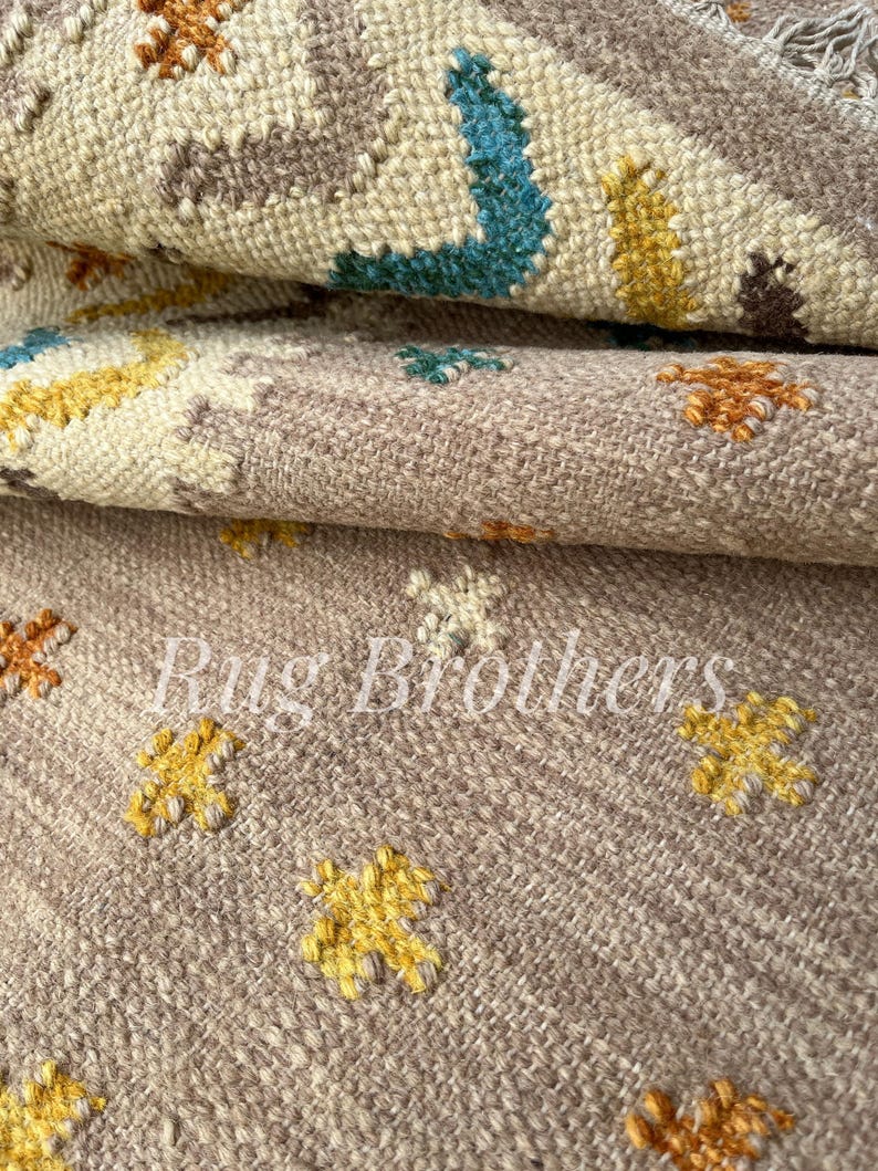 May include: Close-up of a beige and brown woven rug with a repeating floral pattern in yellow, blue, and orange. The rug has a textured surface and the text "Rug Brothers" is visible.